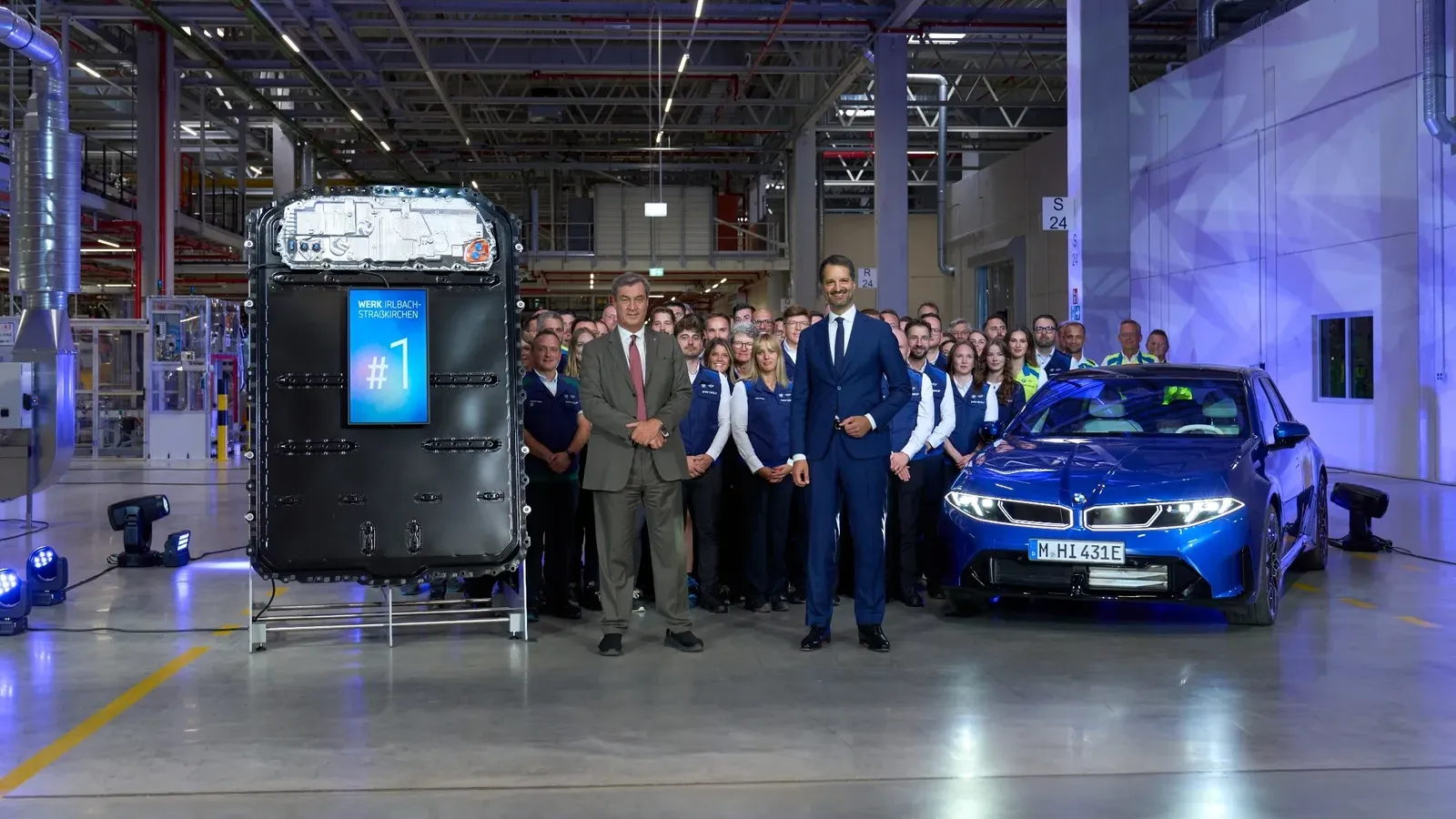 แบตเตอรี่แรงดันสูงที่มีป้าย #1 ตั้งแสดงข้างผู้บริหาร พนักงาน และรถยนต์ BMW ในพิธีเปิดโรงงาน Irlbach-Straßkirchen